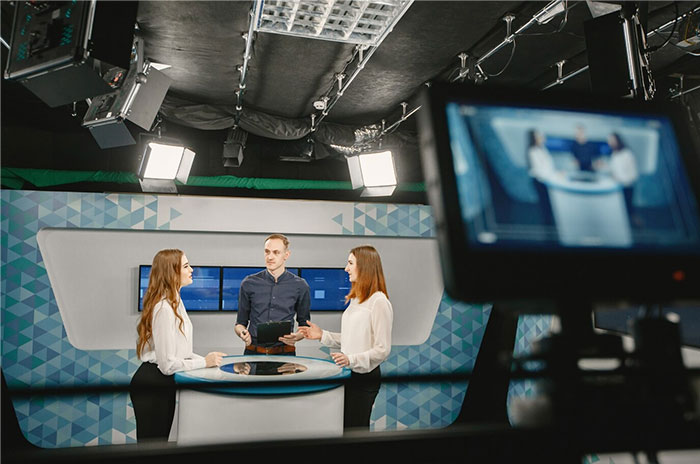 Three people in a studio sharing Hollywood secrets on set, surrounded by cameras and lights.