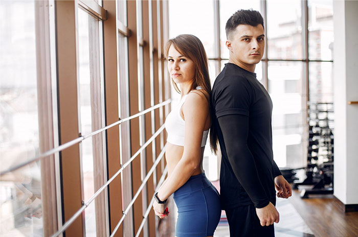 Two people standing back-to-back in a gym, sharing Hollywood secrets with confident expressions.