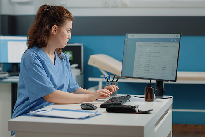 A woman in scrubs works at a desk, possibly doing a job with minimal effort required.