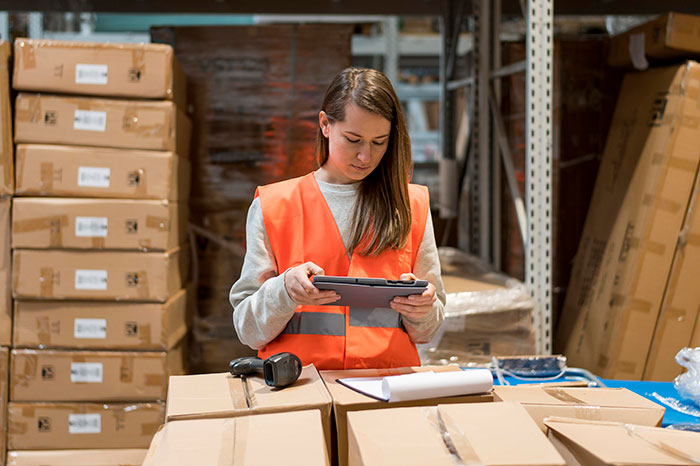 Warehouse worker in orange vest using a tablet, surrounded by boxes, exemplifying low-effort jobs.