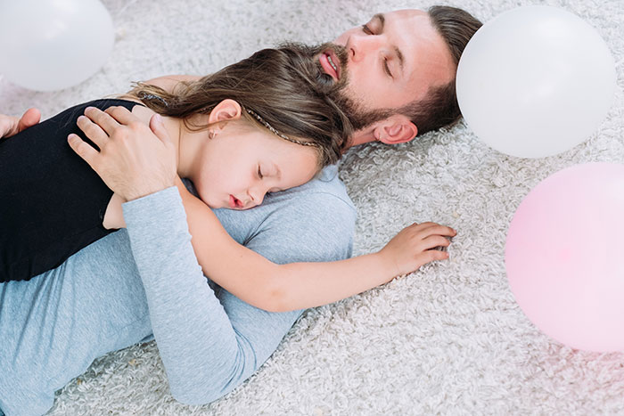 Man and child asleep on a carpet surrounded by balloons, conveying a tender moment in a family setting.