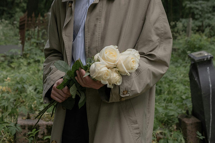 Person in a trench coat holding white roses in a cemetery setting, embodying an embarrassing moment theme.