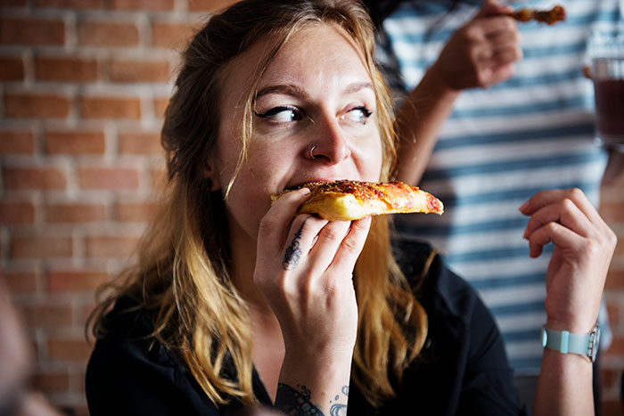 Woman eating pizza, smiling, in a casual setting, capturing a light-hearted embarrassing moment.