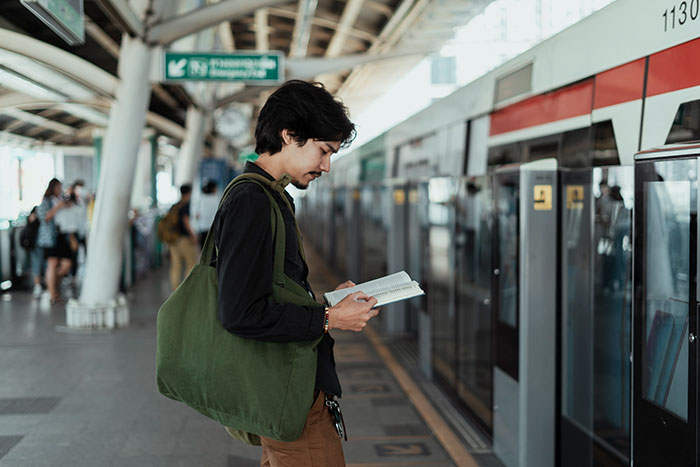 Person reading a book at a train station platform, wearing a green bag, reflecting on embarrassing moments.