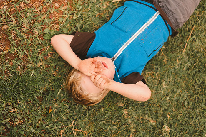 Child in a blue vest covering face, lying on grass, illustrating an embarrassing moment.
