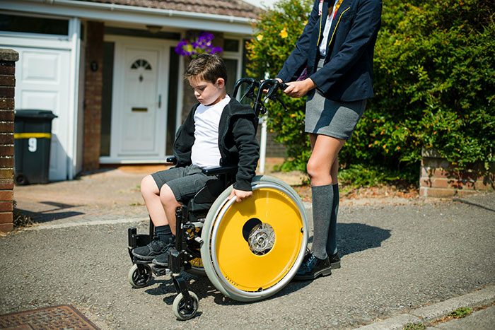 Child in a wheelchair assisted by another child outdoors, reflecting on an embarrassing moment.