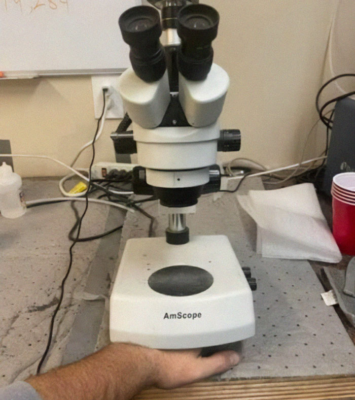 A microscope on a cluttered lab table with someone's hand beneath it; nothing interesting.