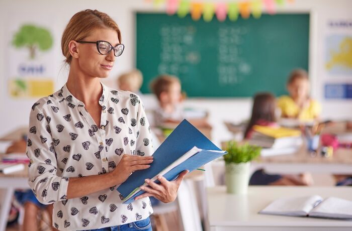 Teacher in classroom holding blue folders, with students in the background, illustrating changing occupations.