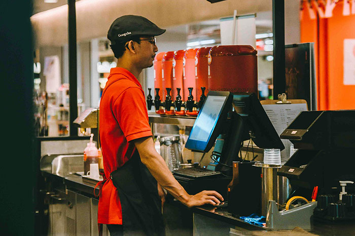 Staff member in red uniform at counter, focusing on screen, in a restaurant setting.