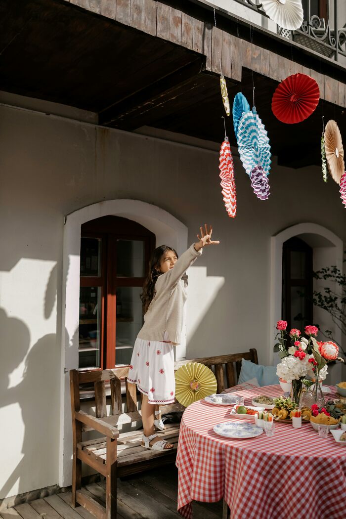 A girl in a cozy setting reaches for hanging decorations by a table with a checkered cloth and flowers, reflecting secret judgments.
