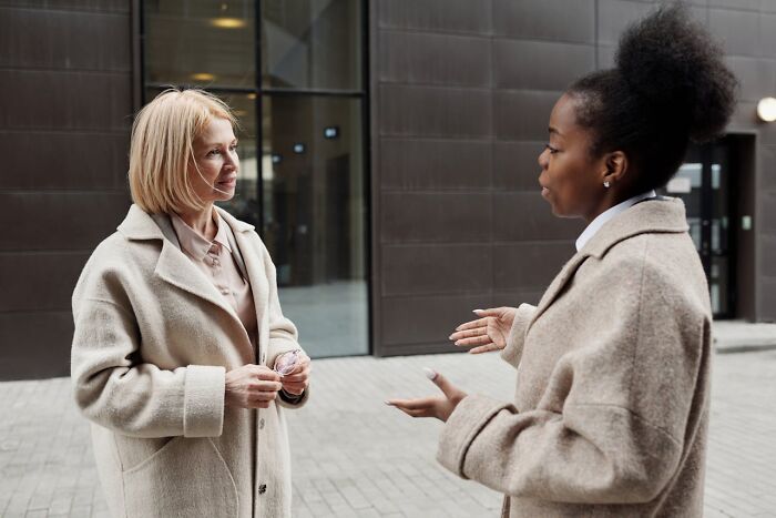 Two women in conversation outside a building, both wearing beige coats and displaying friendly body language.