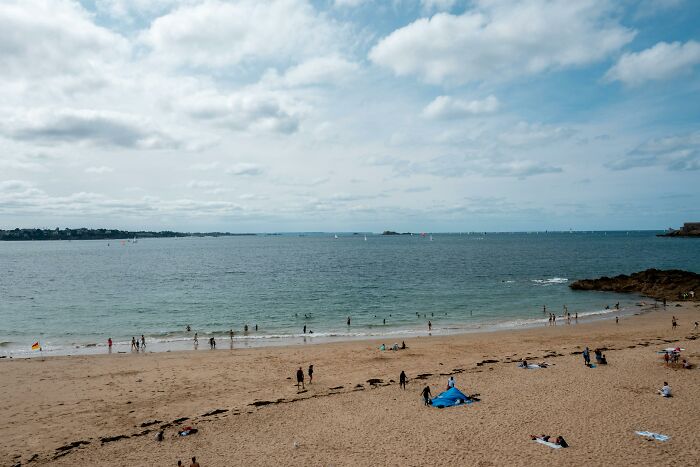 Beach scene with people enjoying a nudist lifestyle, swimming and relaxing under a partly cloudy sky.