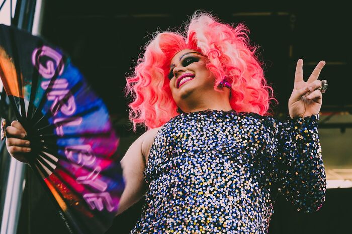 Person in vibrant outfit with pink wig and fan, posing at a lively work Christmas party after potluck.