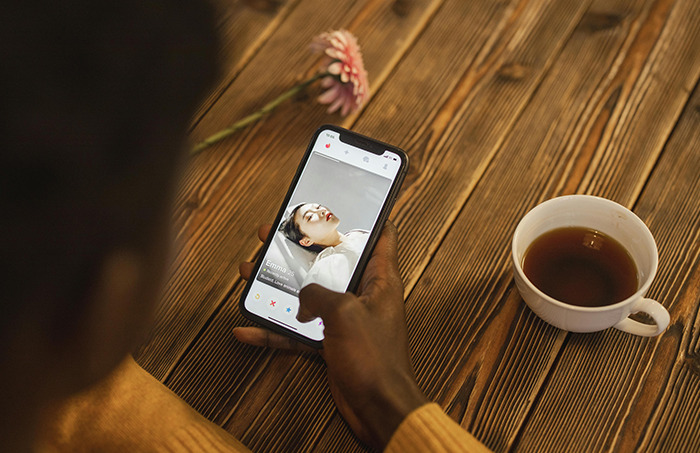 Person swiping on a dating app, illustrating the throning trend, with a flower and tea on a wooden table.