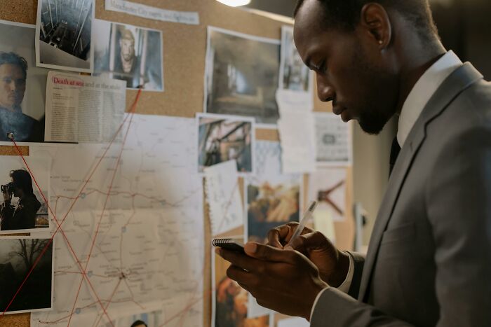 Man in a suit taking notes in front of a pinboard, analyzing clues, representing people knowing secrets.