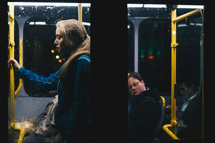 Person in a blue jacket stands on a dimly lit bus, appearing thoughtful about reduced work hours.
