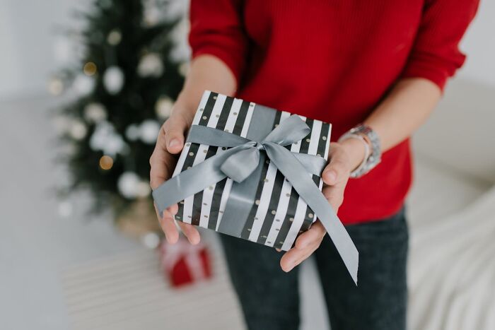 Woman in red sweater holding a striped gift box with a silver ribbon, Christmas tree in the background.