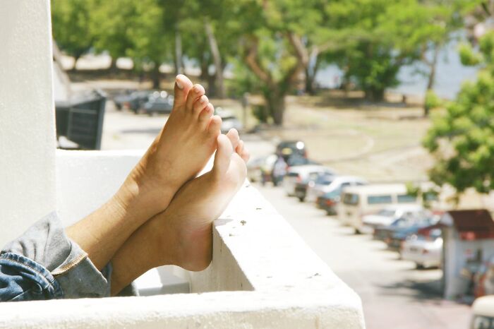Feet resting on a balcony, surrounded by greenery and parked cars, illustrating unusual relaxation.