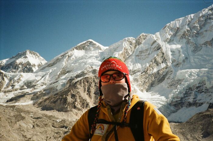 Person in winter gear standing in front of snowy mountains.