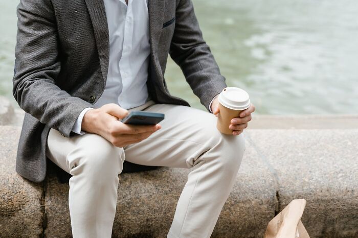 Person sitting on a stone ledge, holding a phone and coffee, exemplifying really messed up moments of multitasking outdoors.