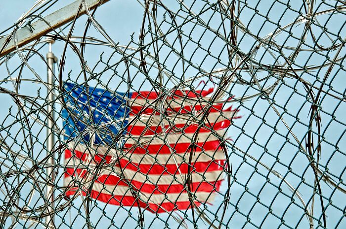 Barbed wire fence with the American flag blurred in the background, symbolizing secrets and barriers.