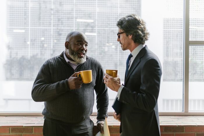 Two colleagues chatting at a work Christmas party, holding coffee mugs near a window.