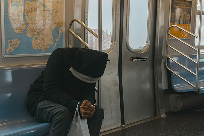 Person in a hoodie sitting on a subway, face hidden, holding a bag.