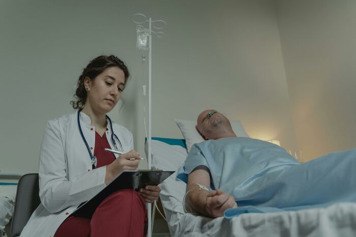 A female doctor in a hospital room, noting details while sitting beside a patient lying in bed, showcasing WomenInMaleFields.