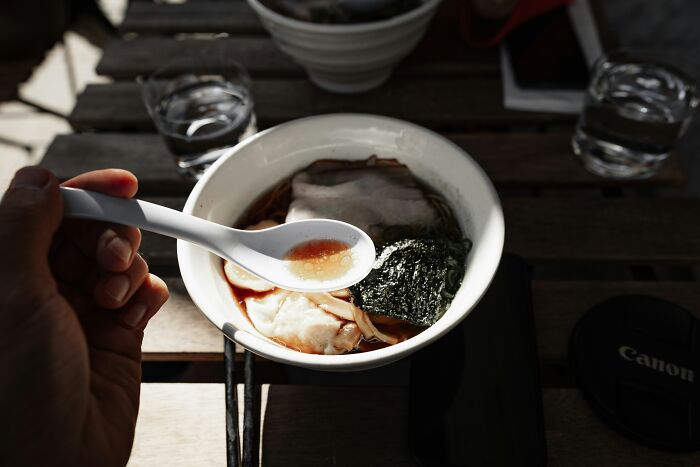 A person holding a spoon over a bowl of ramen on a wooden table, embodying frugal home dining.