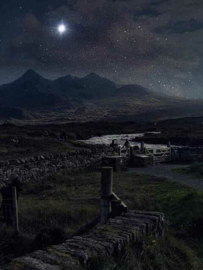 Starry night sky over Scottish landscape with distant mountains and a stone fence in the foreground.