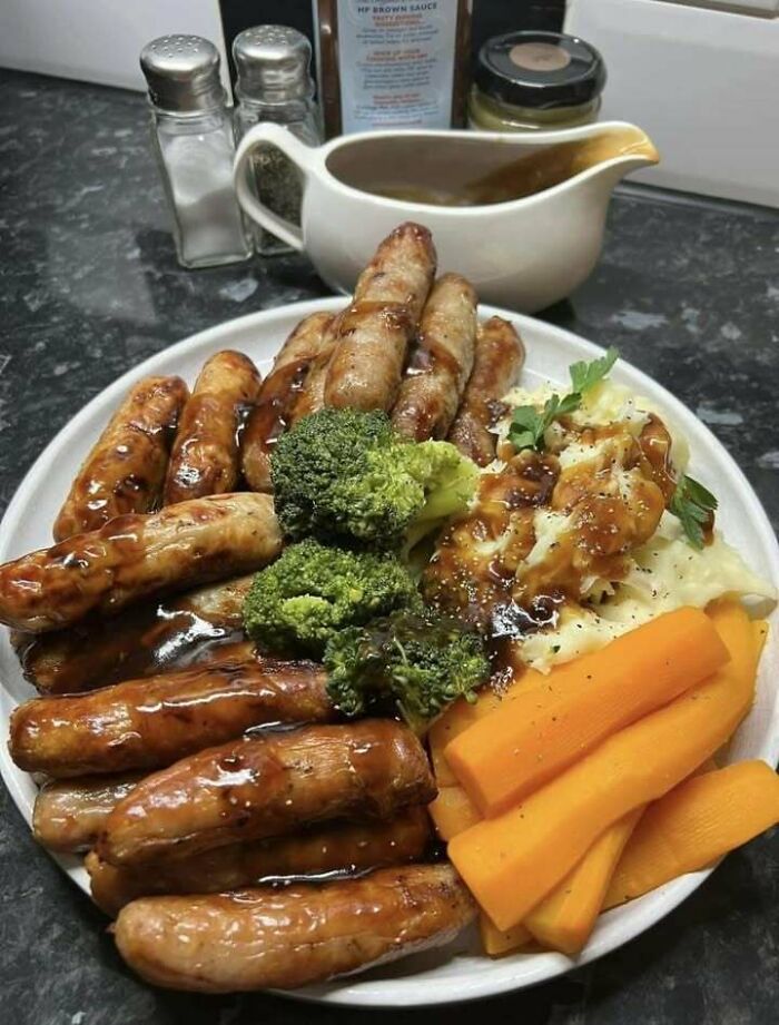 Scottish meal with sausages, broccoli, mashed potatoes, and carrots, served with gravy and condiments in a kitchen.
