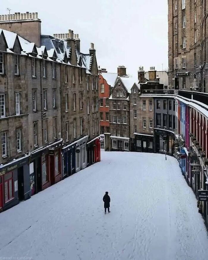 Snow-covered Edinburgh street with historic buildings, highlighting Scotland's charm and humor with a lone figure walking.