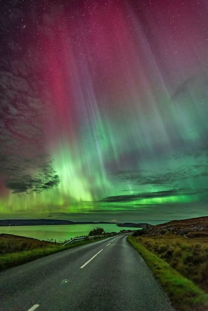 Aurora borealis illuminating a rural road in Scotland under a vibrant night sky.