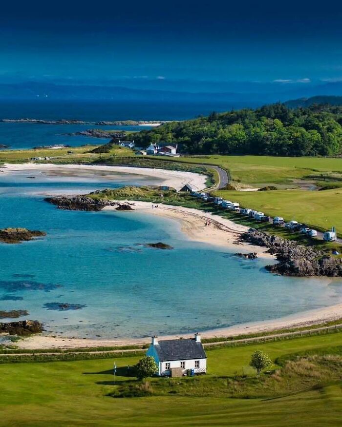 Scottish coastal landscape with a white cottage, blue sea, and lush greenery under a clear sky.
