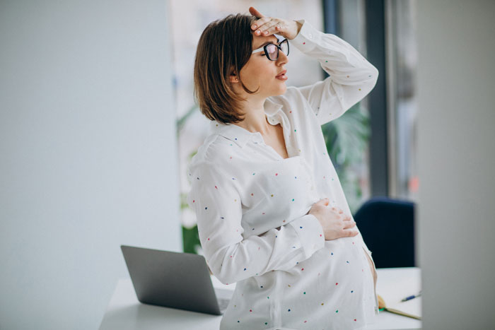 Pregnant woman in white shirt, holding her head, standing near a laptop, appearing concerned.