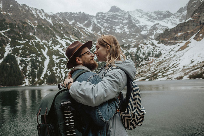 A couple in winter clothing stands by a snowy mountain lake, embracing each other with backpacks.