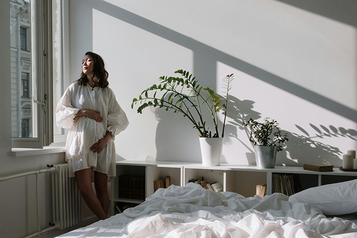 Pregnant woman in a serene bedroom, standing by the window with light streaming in and plants in the background.