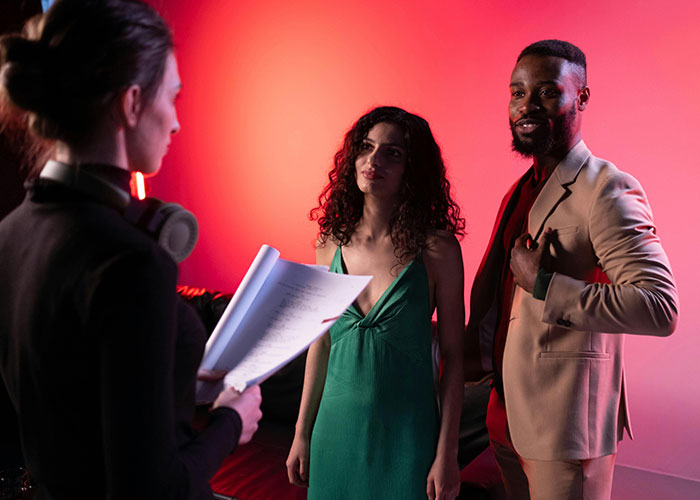 Reality TV show cast discussing script on set with a director, under dramatic red lighting.