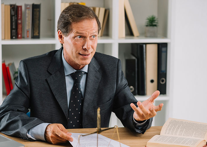 Man in a suit speaking at a desk, surrounded by books, sharing insights on reality TV shows.