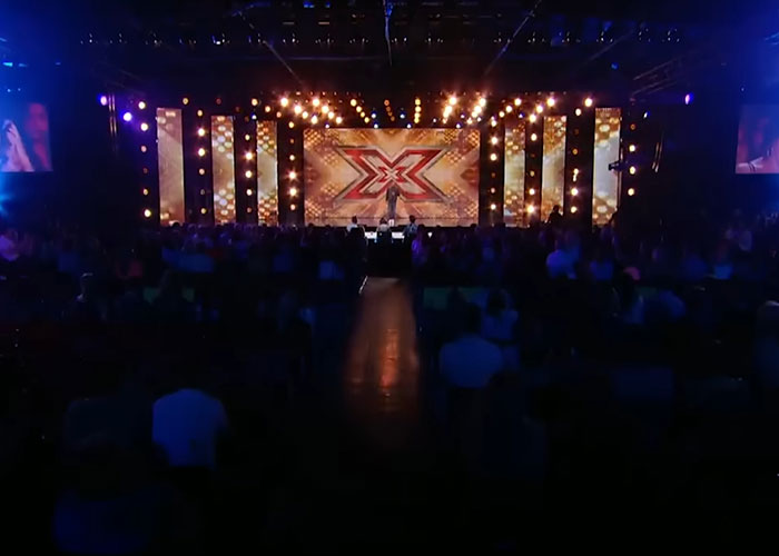 Reality TV show stage with vibrant lighting and audience, featuring an illuminated 'X' symbol.