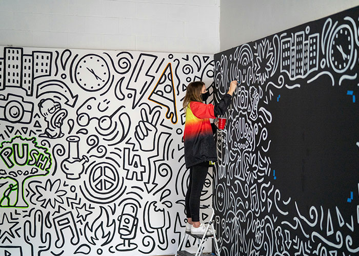 Person on a ladder drawing intricate reality TV-inspired doodles on a wall.