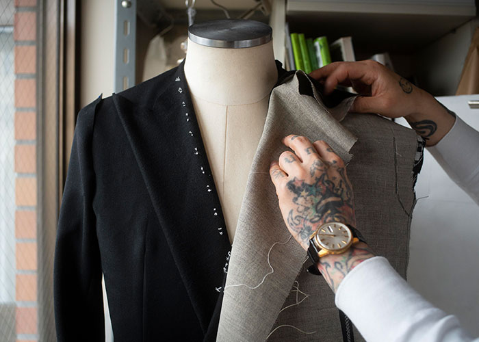 Tattooed tailor adjusting a jacket on a mannequin, offering insight into reality TV fashion design.