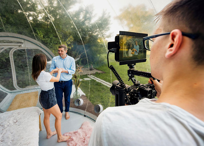 Filming a reality TV scene in a transparent dome, featuring a couple and a cameraman capturing the moment.