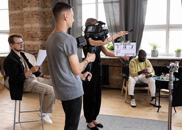 Crew filming a reality TV show scene with a clapperboard, camera operator, and two seated participants on set.
