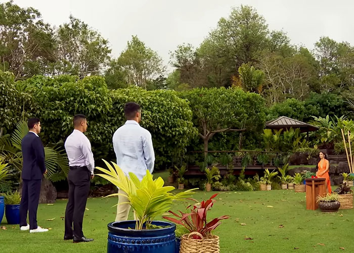 Reality TV scene with three men in a garden facing a woman seated at a wooden table.