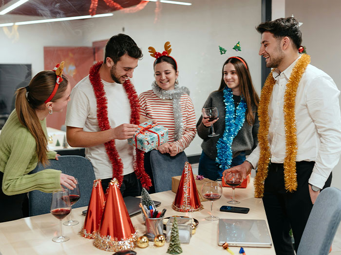 Office Christmas party with people in festive accessories, exchanging gifts and holding drinks around a decorated table.