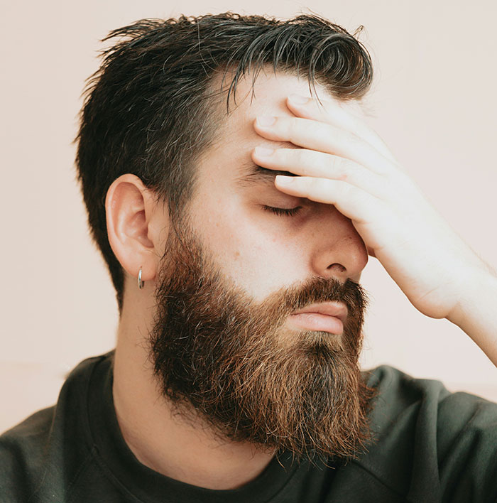 Man with beard looking stressed, hand on forehead, illustrating an awkward living situation involving his girlfriend moving in.