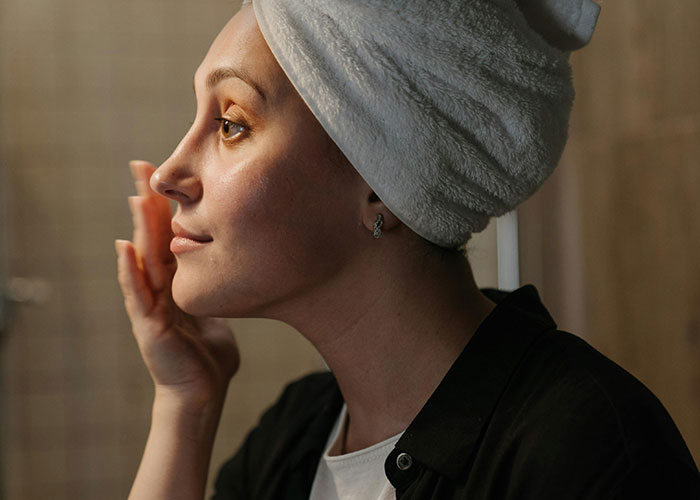 Person applying makeup with a white towel on their head in a bathroom setting.