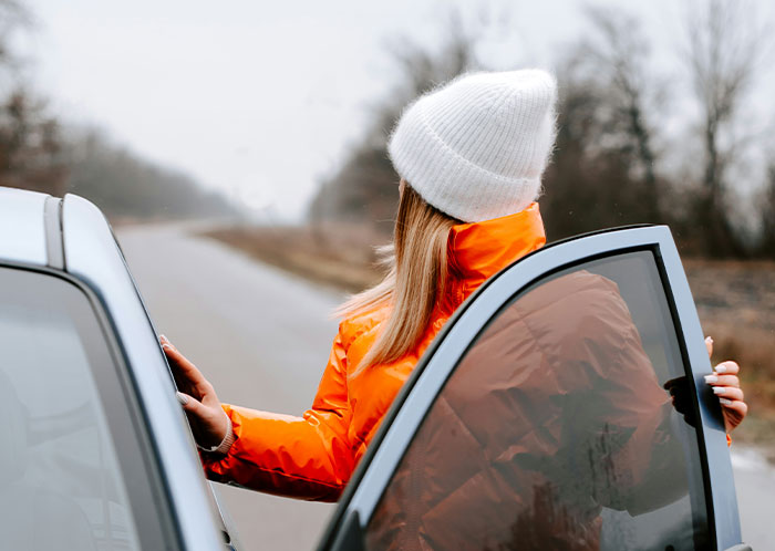 Person in orange jacket and white hat looking out of car door at night road.