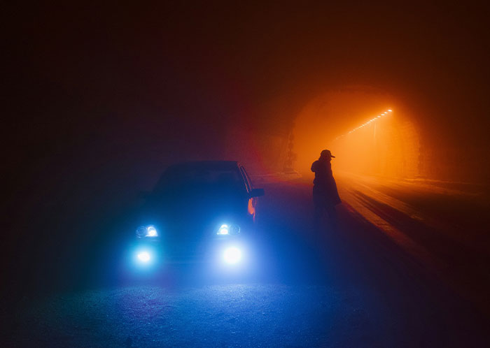 Car with bright headlights in foggy tunnel at night, silhouette of a person nearby. Weirdest night driving scene.